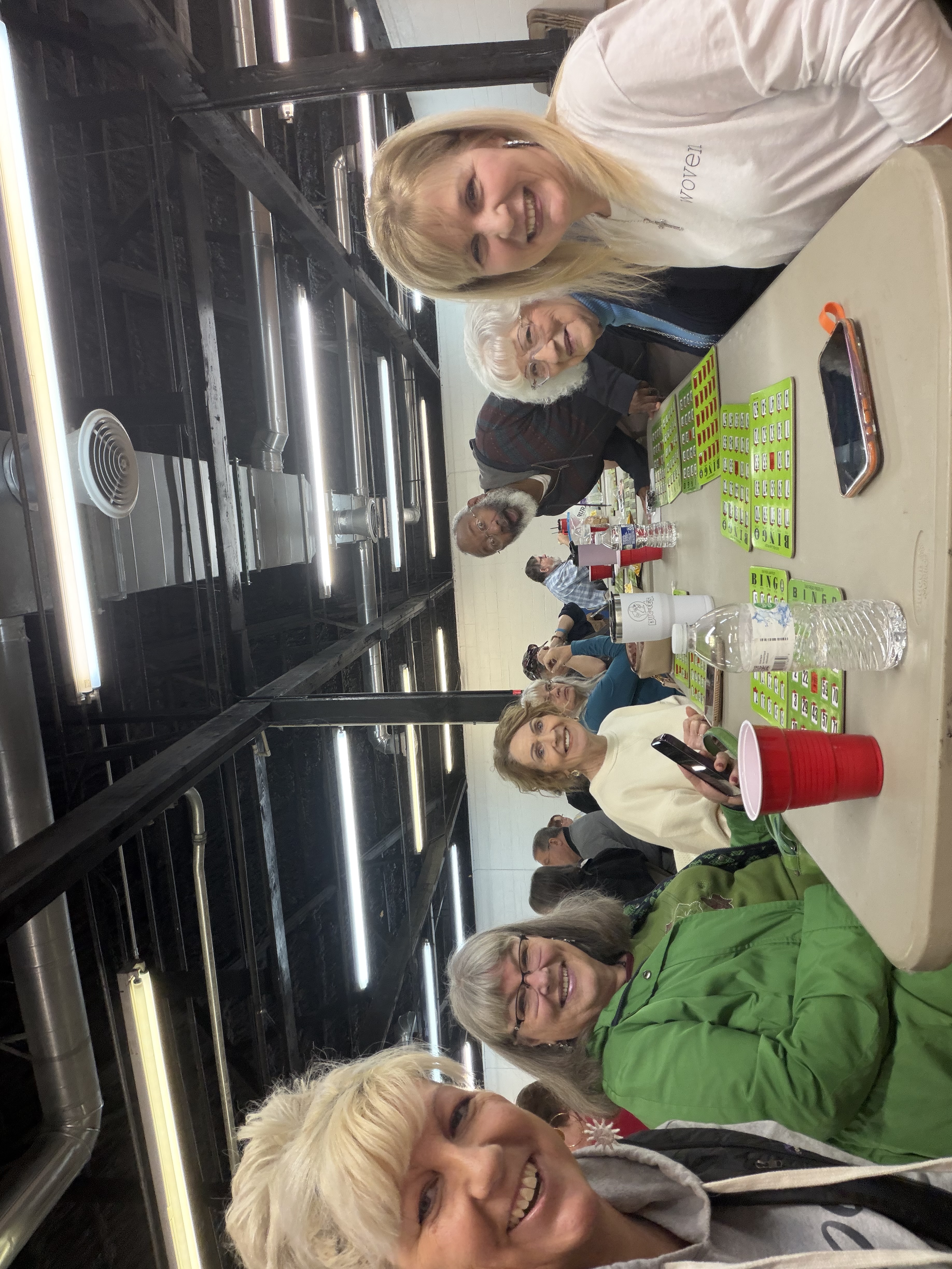 Six smiling friends gathered around a table covered in green bingo cards at a community bingo night