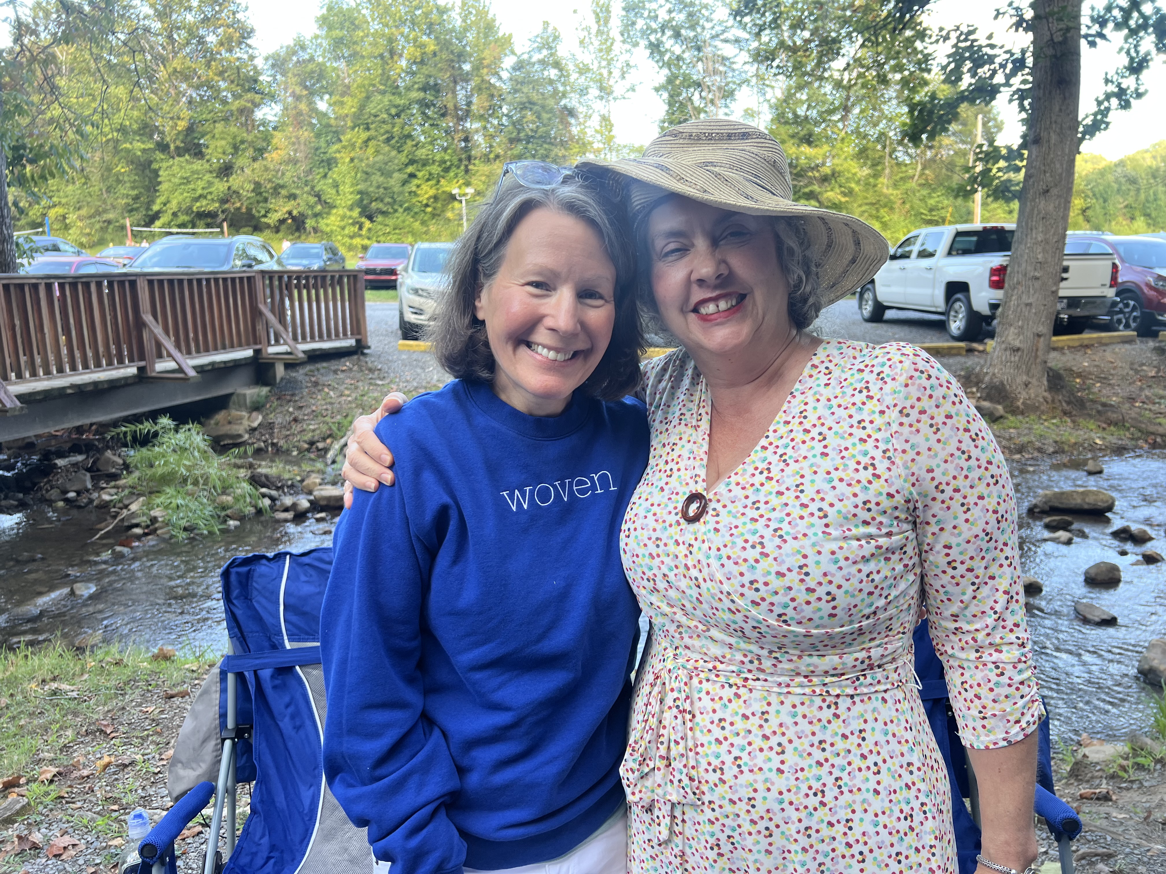 Two women smiling arm in arm beside a creek in the woods on a sunny afternoon