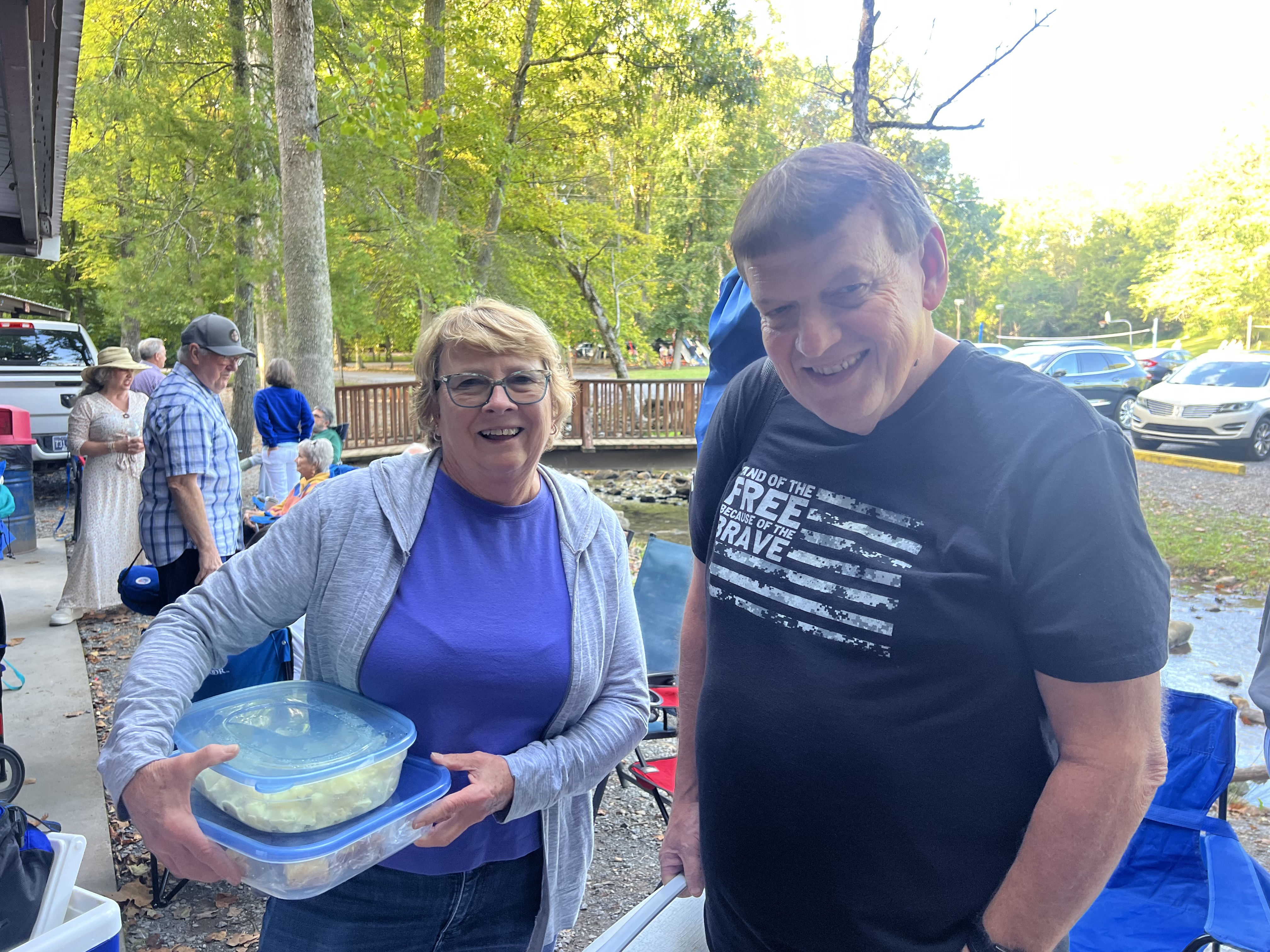 A couple smiling at a creekside picnic, holding a covered container of potato salad to share with the group